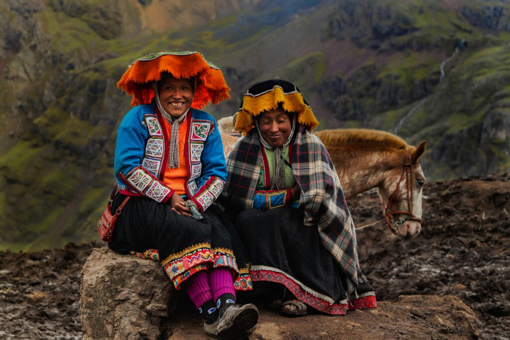 Two women in traditional Andean clothing sit on a rock in the Peruvian mountains, accompanied by a horse.